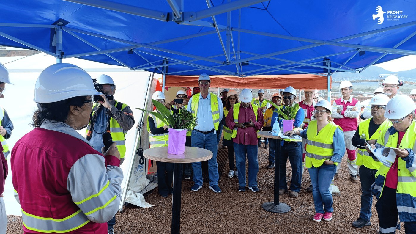Cérémonie d'inauguration sur le site minier de Prony Resources à Goro : des invités en casque et gilet haute visibilité réunis sous un barnum bleu, une plante en pot sur une mange-debout, installations industrielles en arrière-plan.